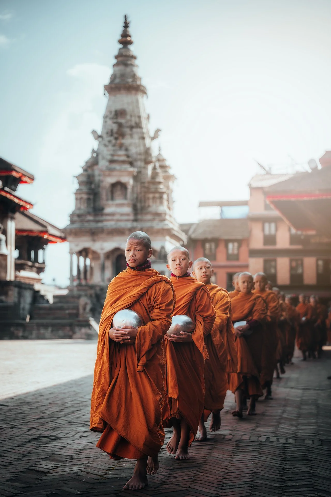 Monaci buddisti in tunica arancione che camminano in fila con ciotole in mano davanti a un tempio in stile nepalese.