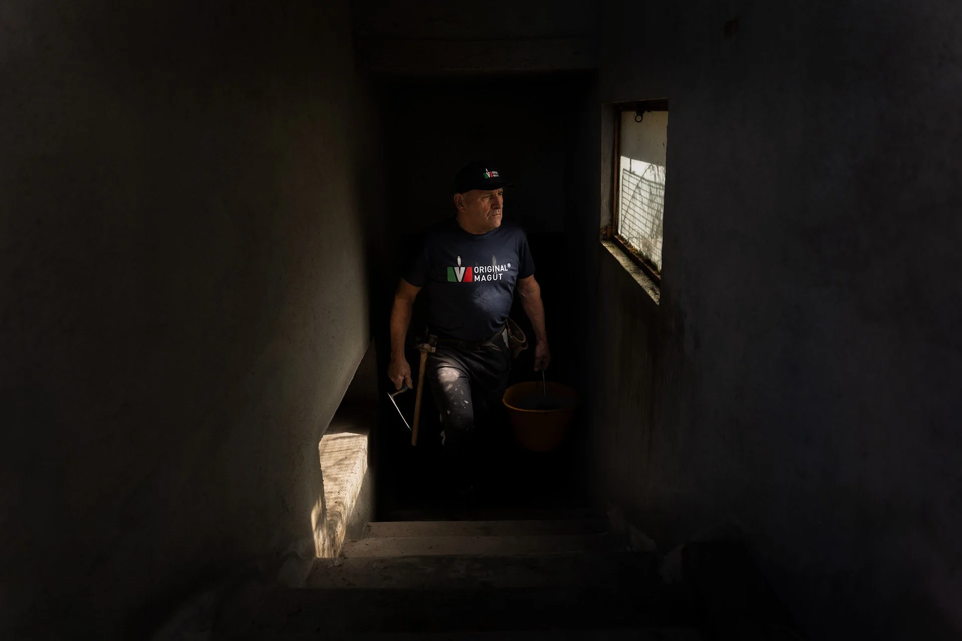 Uomo con cappellino nero e maglietta con logo 'Original Magùt' cammina in un'area buia con una finestra luminosa a destra, portando un secchio e un attrezzo