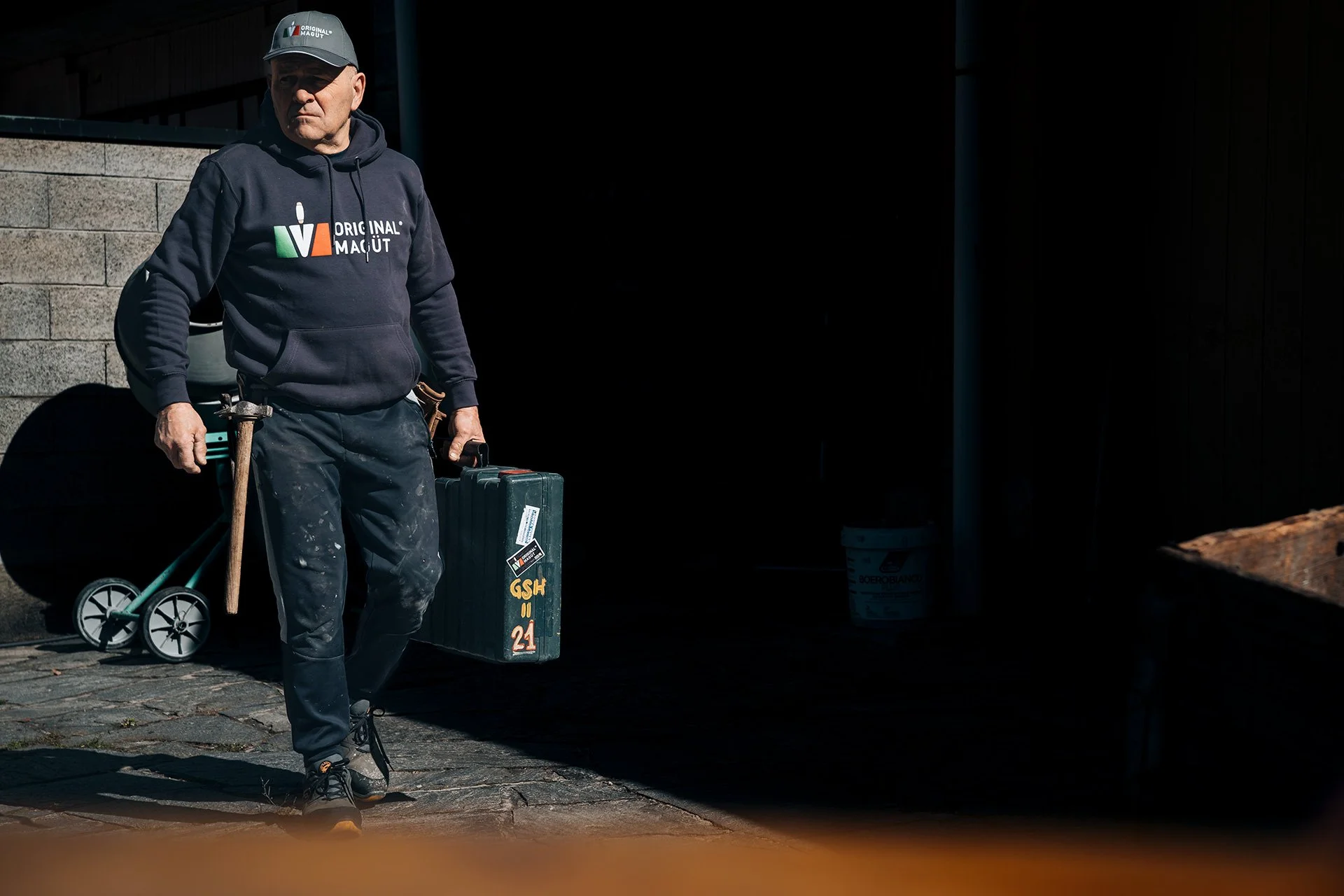 Uomo anziano che cammina, indossa felpa con logo 'Original Magut' e cappello, tiene un attrezzo da lavoro e una valigia, sfondo di una parete e di una zona coperta, in scena una giornata di lavoro.