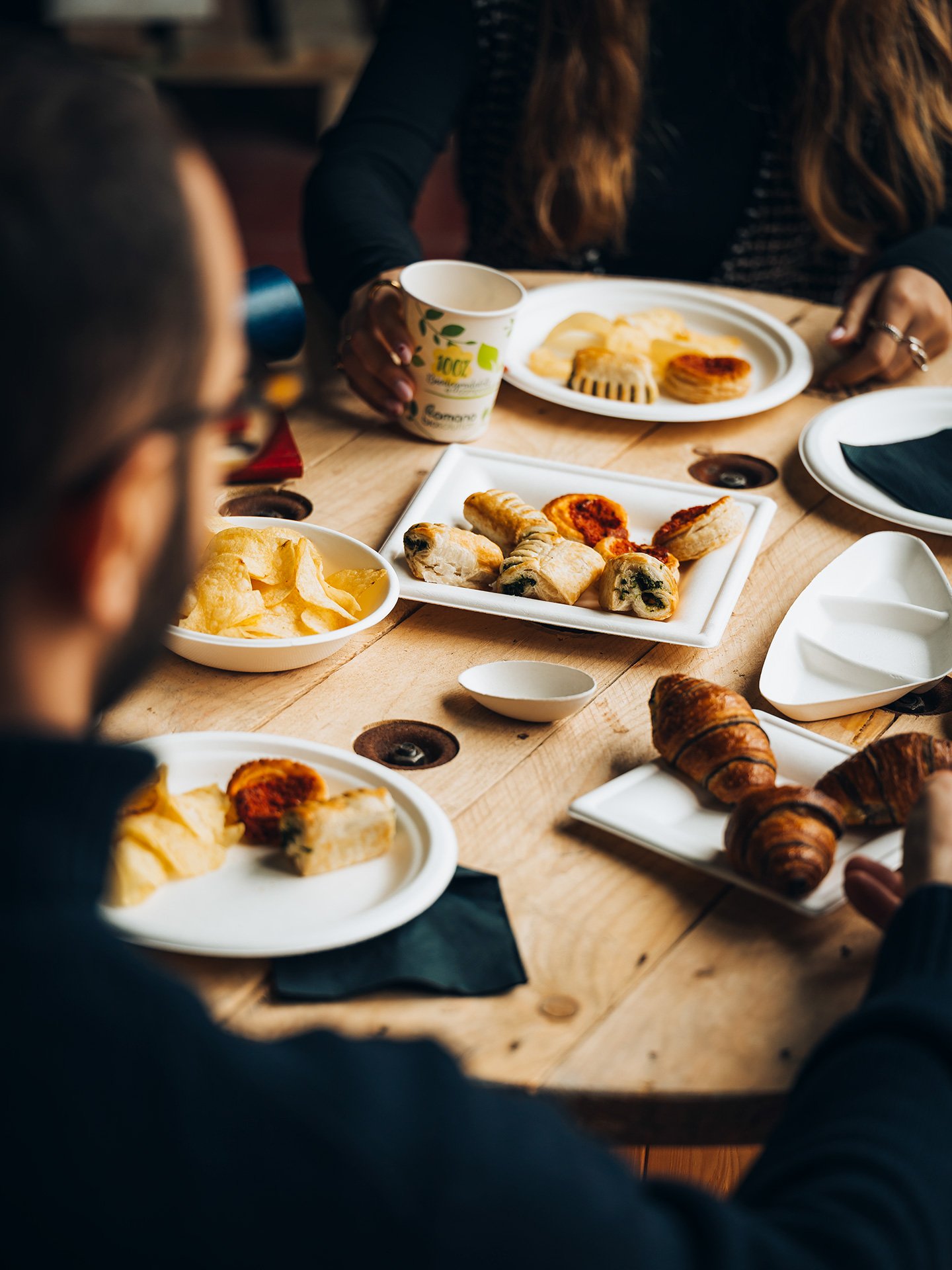 Persone che mangiano una colazione con croissant, pizza, patatine e bevande al tavolo.