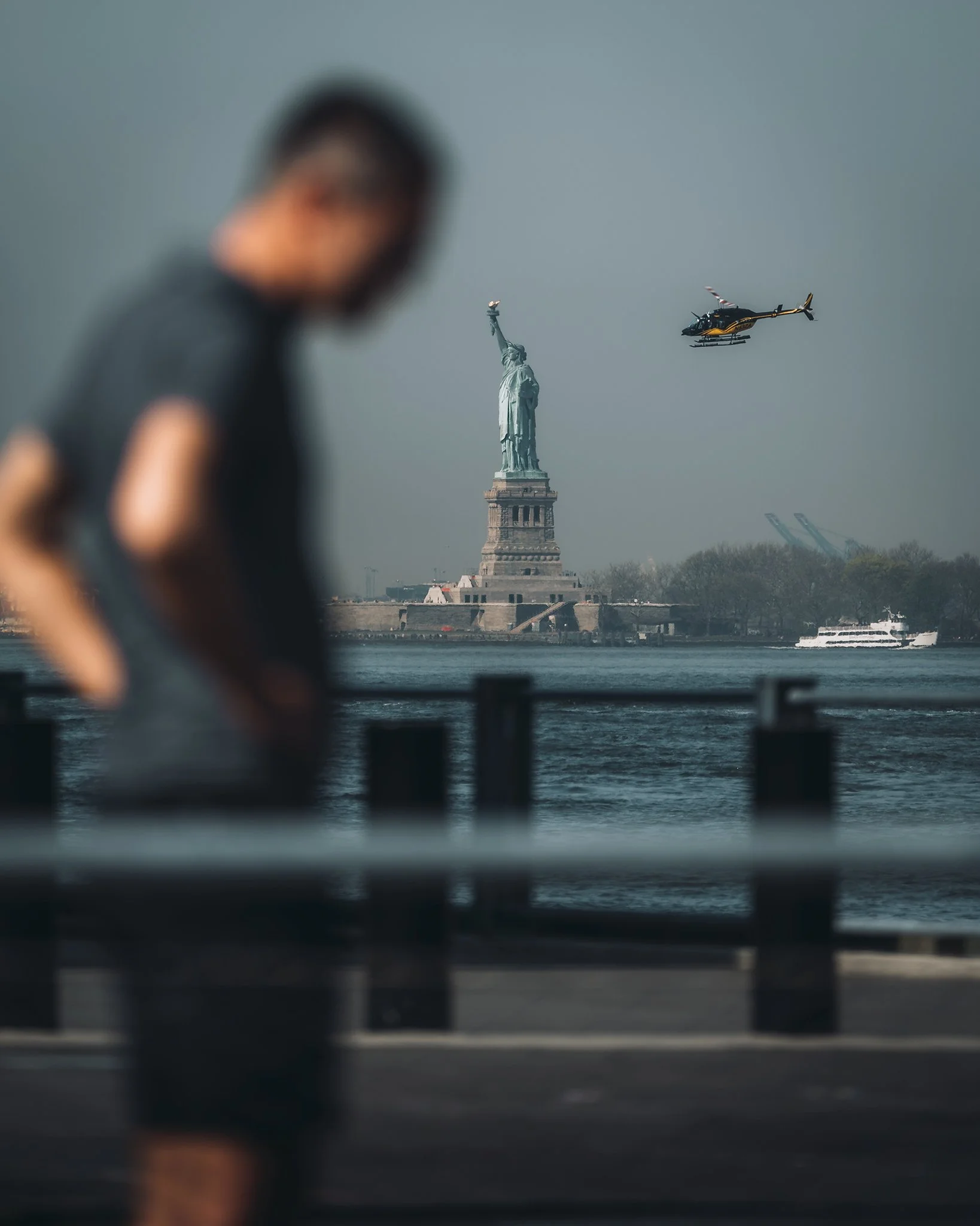 Immagine di una persona sfocata davanti, con la Statua della Libertà e un elicottero sullo sfondo a New York.