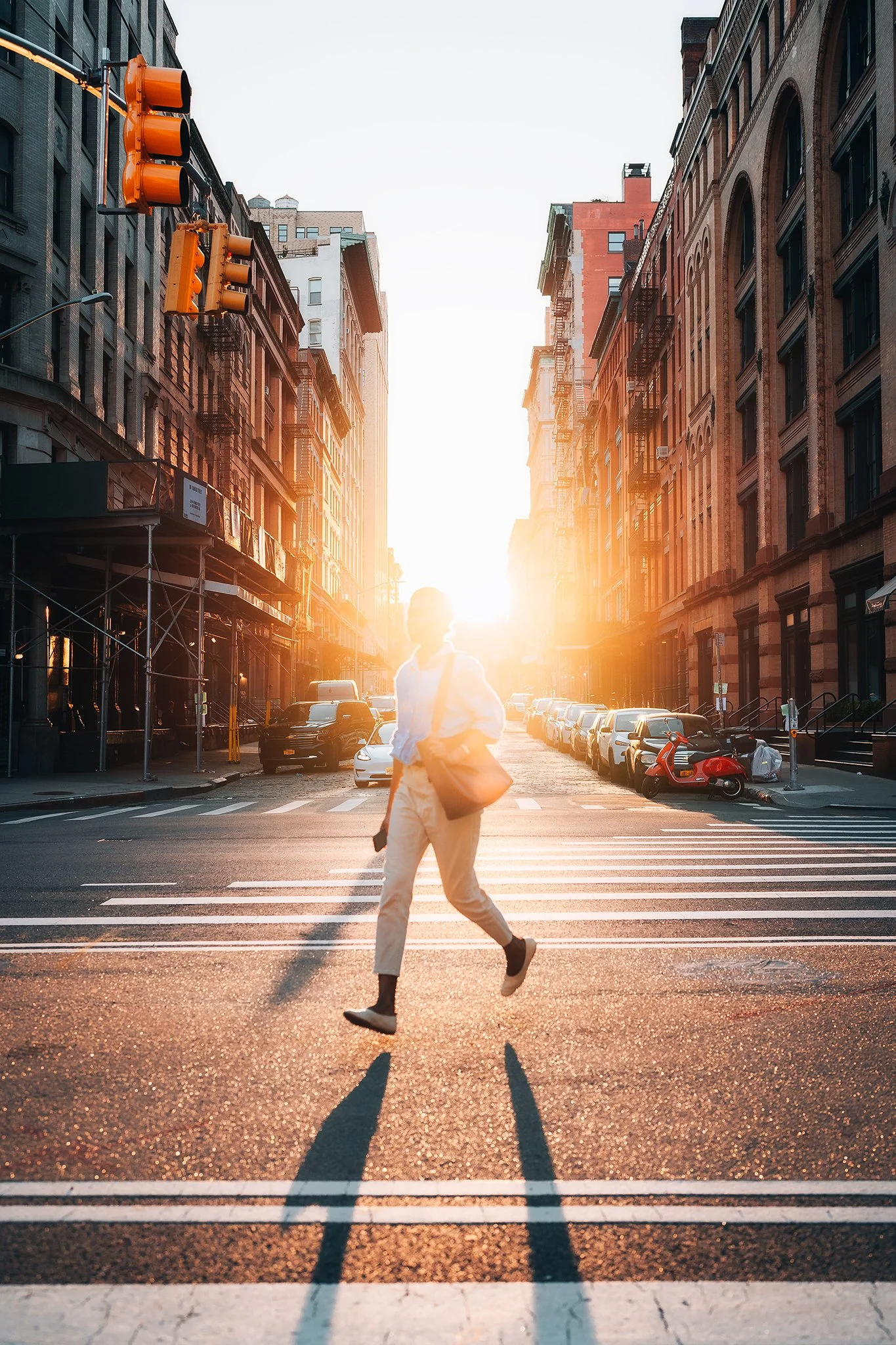 Uomo che attraversa una strada con il sole al tramonto dietro di lui, creando un'ombra lunga, in un quartiere urbano con edifici alti e automobili parcheggiate.