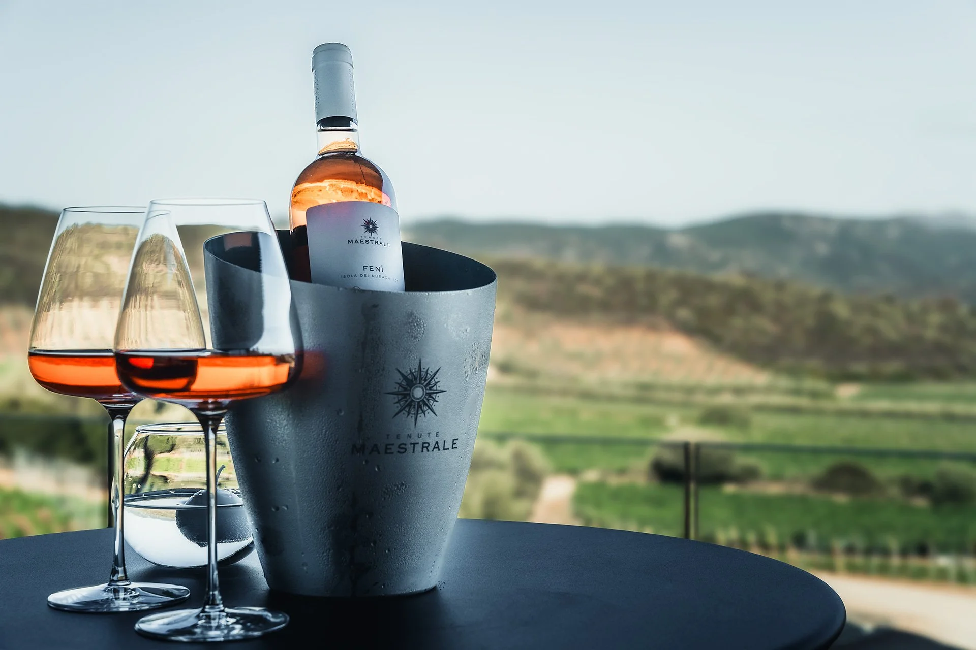 Bottiglia di vino rosato in un secchiello con due calici di vino rosato sul tavolo, con uno sfondo di colline e campi in campagna.