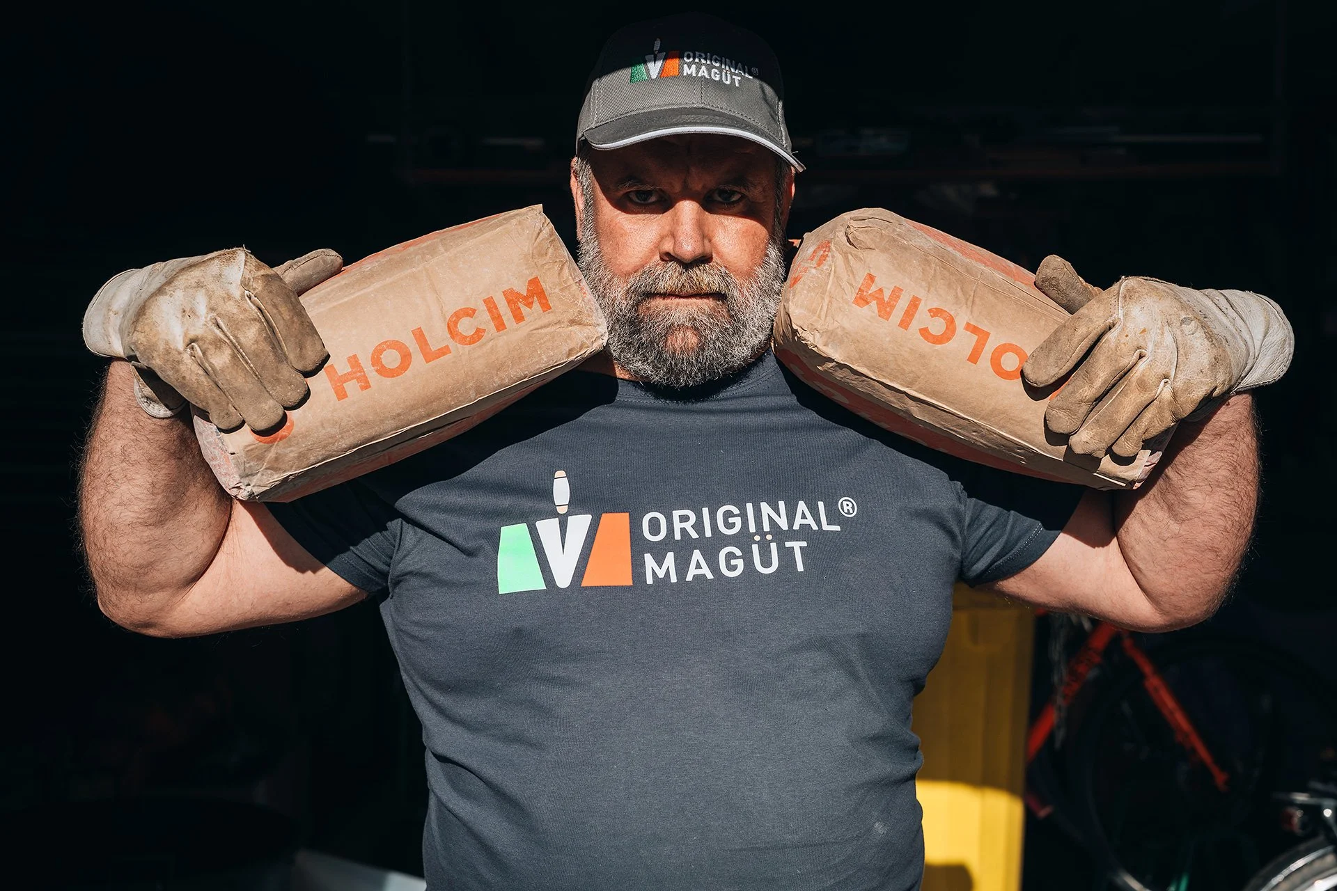 Uomo muscoloso con barba grigia indossa una maglietta con il logo 'Original Magût' e una copricapo con lo stesso logo, porta due sacchi di cemento sulla spalla, indossa guanti da lavoro e guarda verso la fotocamera.