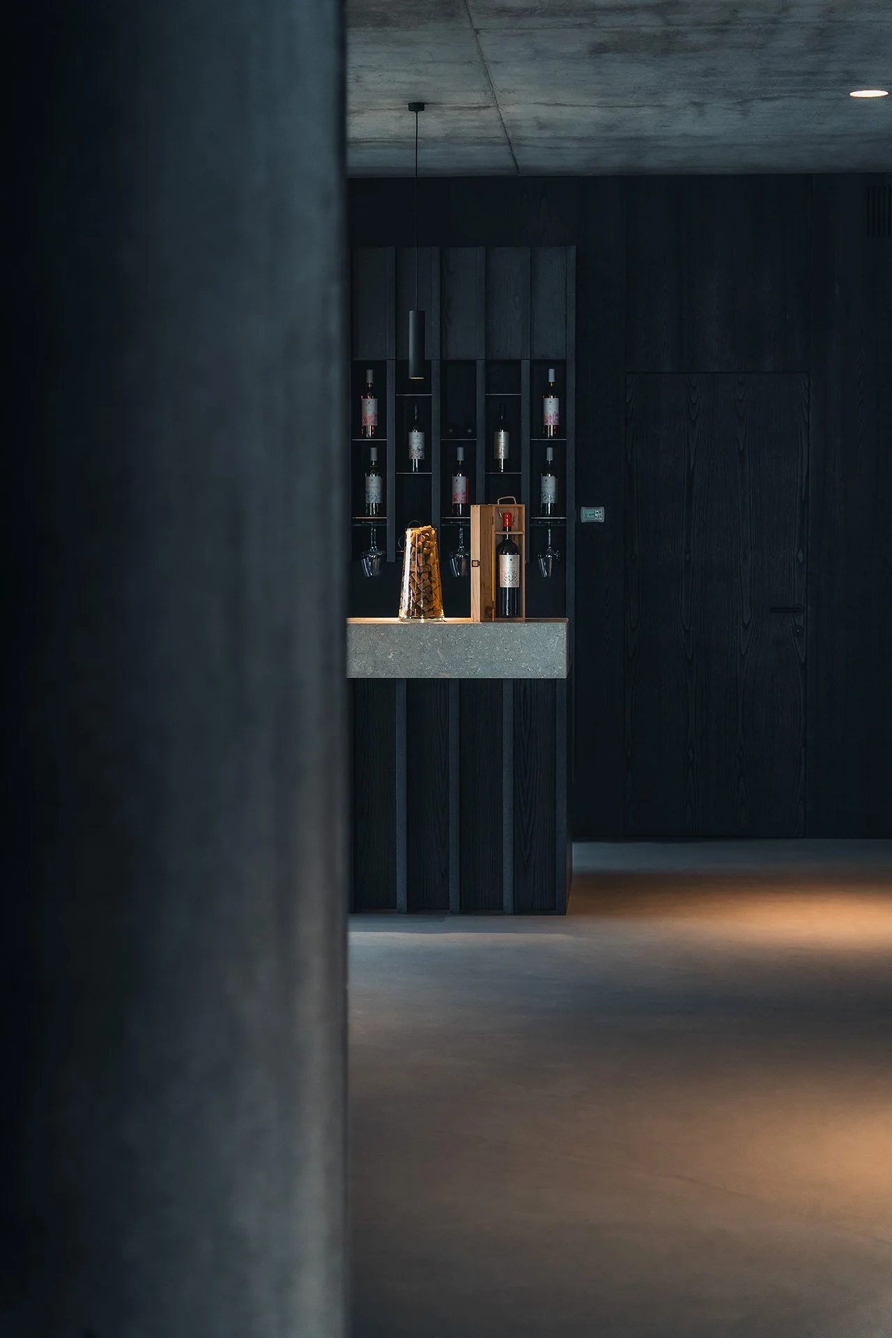 Interno di un ambiente moderno con scaffali neri e bottiglie di vino, illuminato da luci soffuse.
