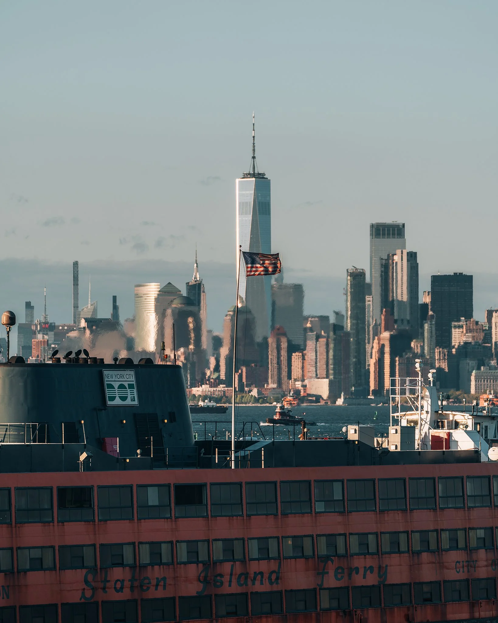 Veduta dello skyline di New York City con il One World Trade Center al centro e un'onda di fumo visibile nel basso. Sullo sfondo, il fiume e alcuni edifici. In primo piano, una nave con bandiera degli Stati Uniti e un cartello con scritto 'Staten Isl
