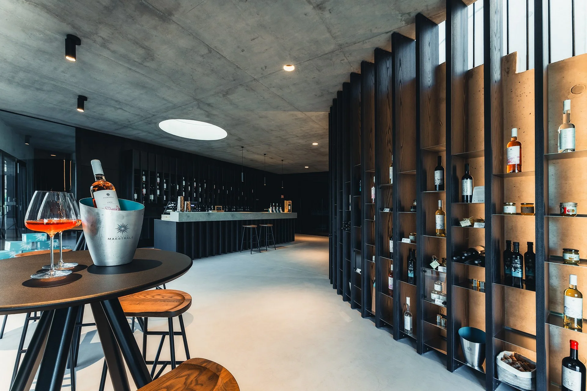 Interno di una moderna enoteca o enoteca di vini con scaffali di bottiglie di vino su una parete di legno, tavoli con bicchieri di vino rosato e un ambiente luminoso con soffitto in cemento e grandi finestre.