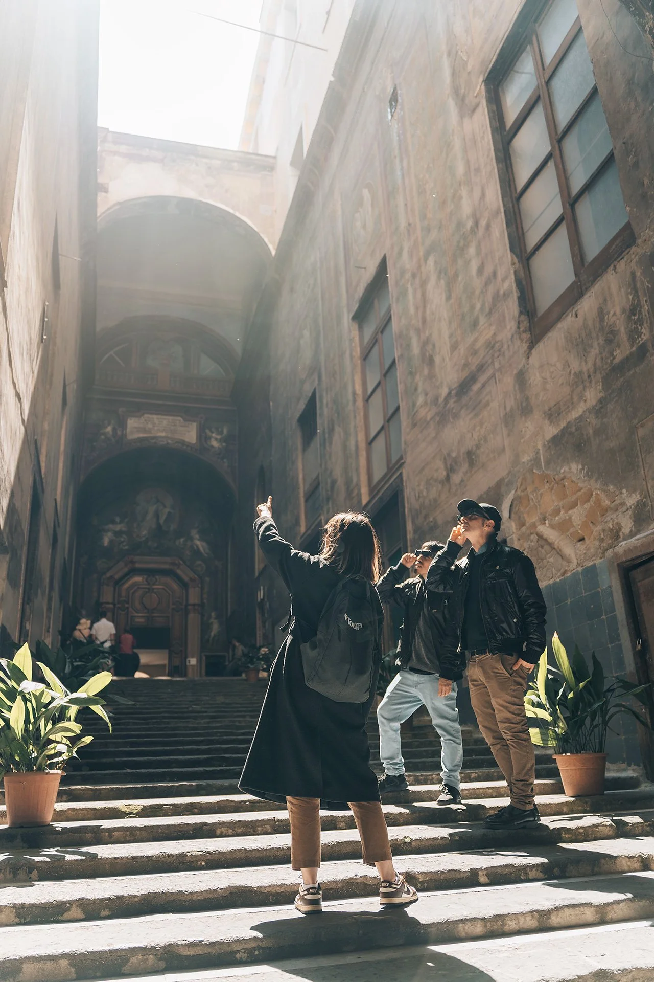 Tre giovani che si incontrano davanti a un ingresso storico con scalinata, all'interno di un edificio antico con pareti in pietra e decorazioni sopra l'entrata.