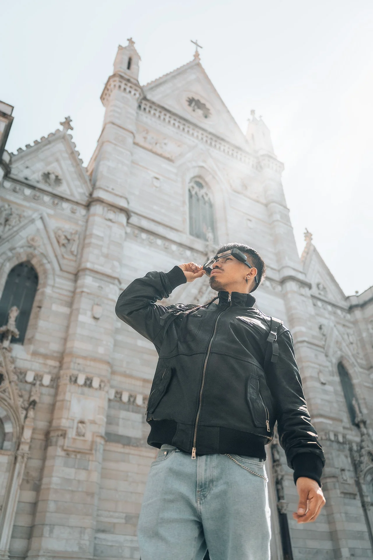 Un uomo con occhiali da sole e giacca nera si trova davanti a una chiesa storica con architettura gotica, in un ambiente urbano.