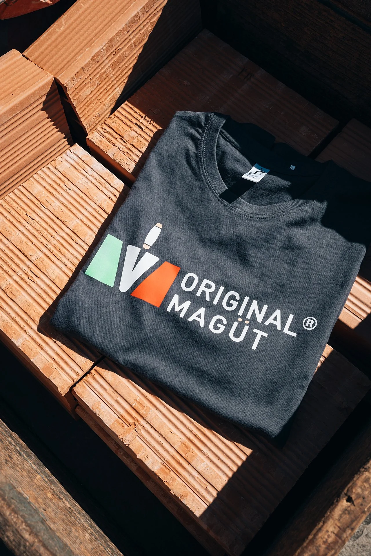 Una maglietta nera con la scritta 'Original Magüt' e un logo colorato, poggiata su un bancale di legno.