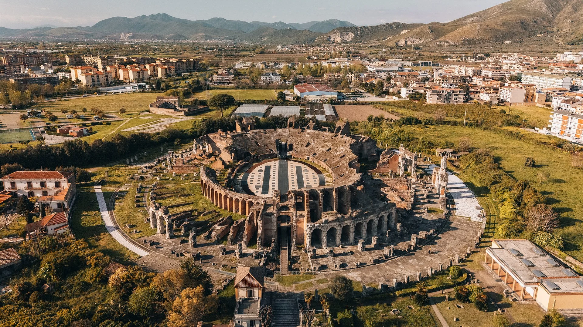 Immagine aerea di un'antica arena romana circondata da un'area verde con edifici moderni e montagne sullo sfondo.