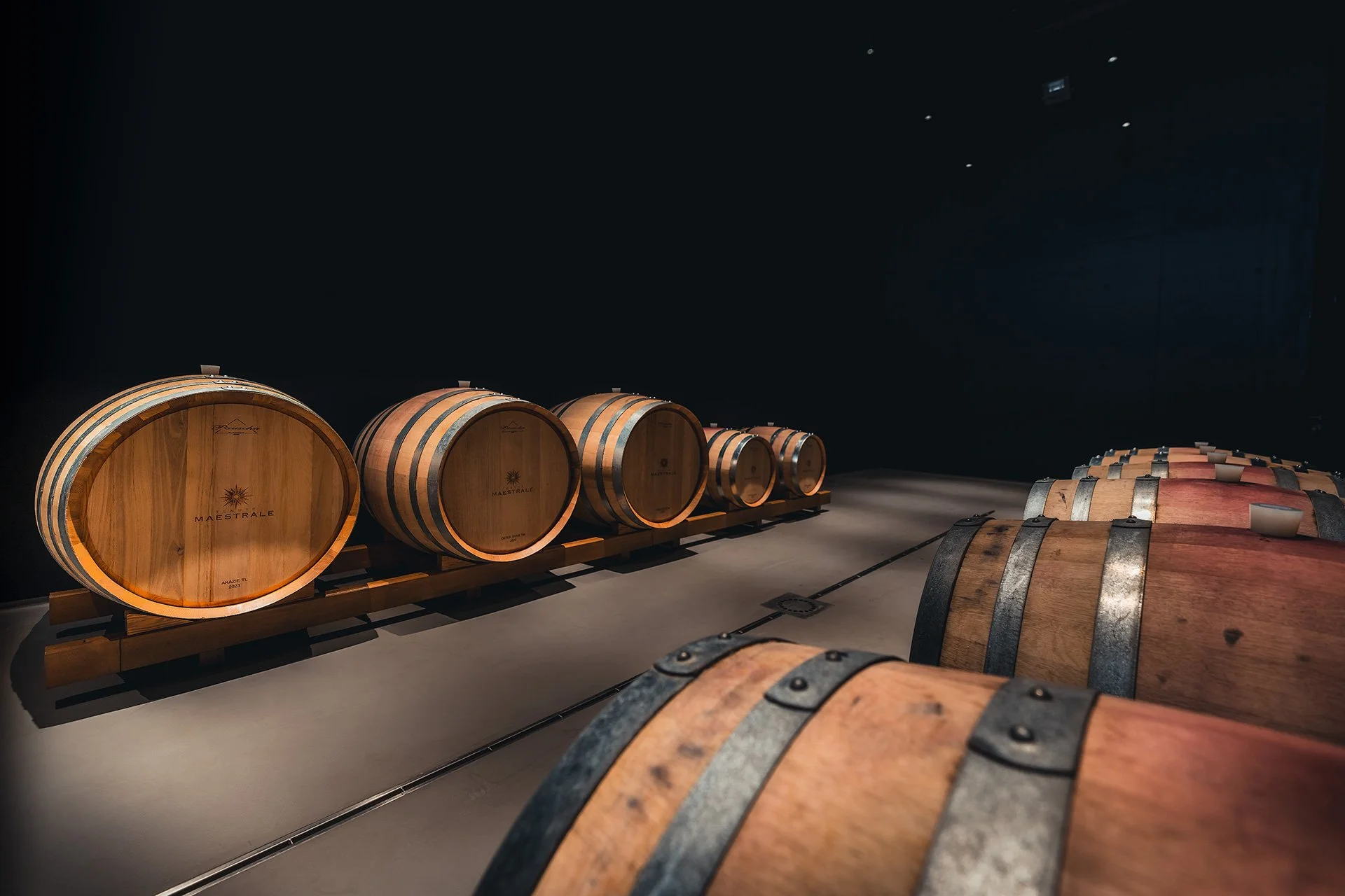 Fate in legno e metallo di botti di vino in una cantina Al centro, botti di vino di dimensioni variabili disposte in fila, con il marchio 'Maestrale' e la parola 'Ancora' sul lato frontale. Sul lato destro, altre botti più piccole sono posizionate in