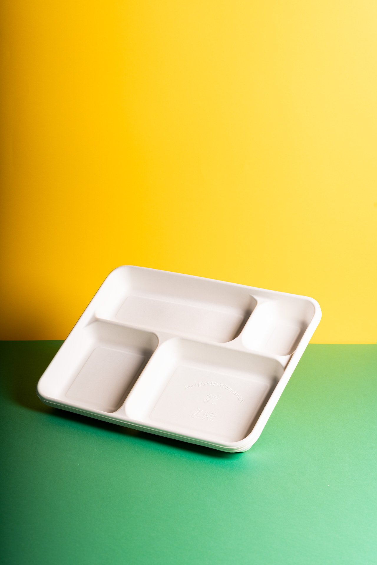 Piatto bianco per alimenti biodegradabile, diviso in quattro scomparti, posato su uno sfondo verde e giallo