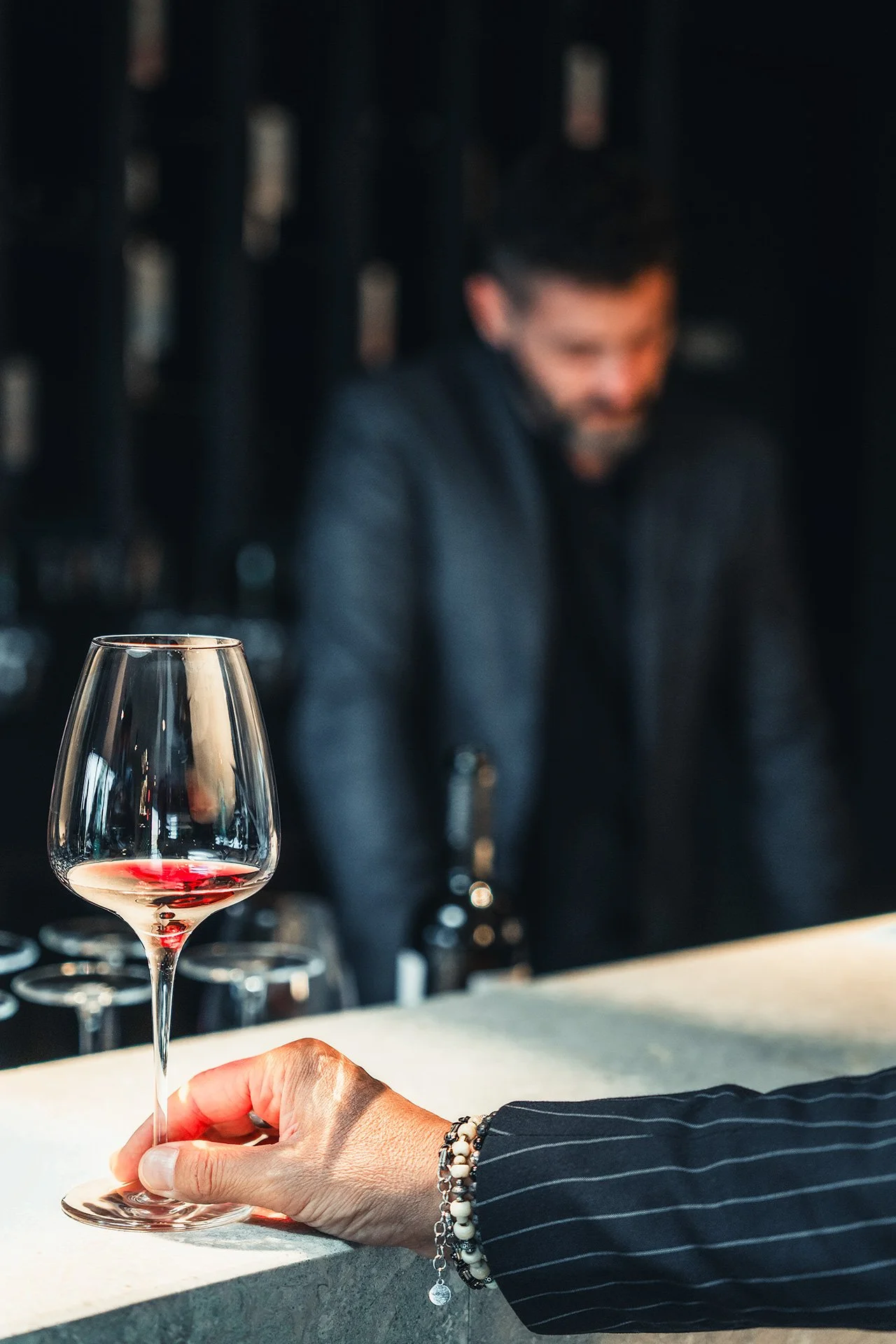 Una mano tiene un bicchiere di vino rosso, con uno sfondo di un bar o ristorante con una persona che sorride e sfocata.