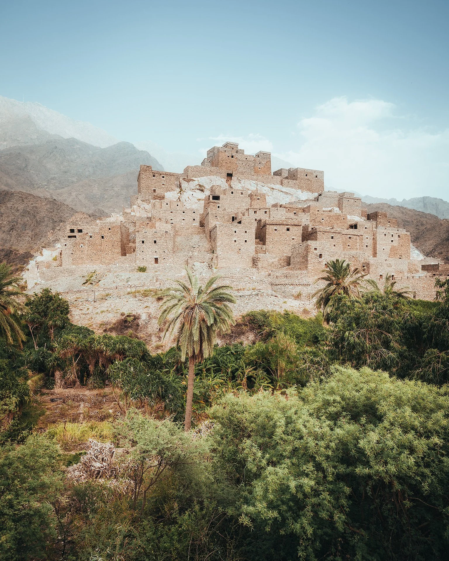 Una fortezza antica in pietra costruita su una collina rocciosa con alcuni alberi di palma in primo piano e montagne sullo sfondo, sotto un cielo chiaro.