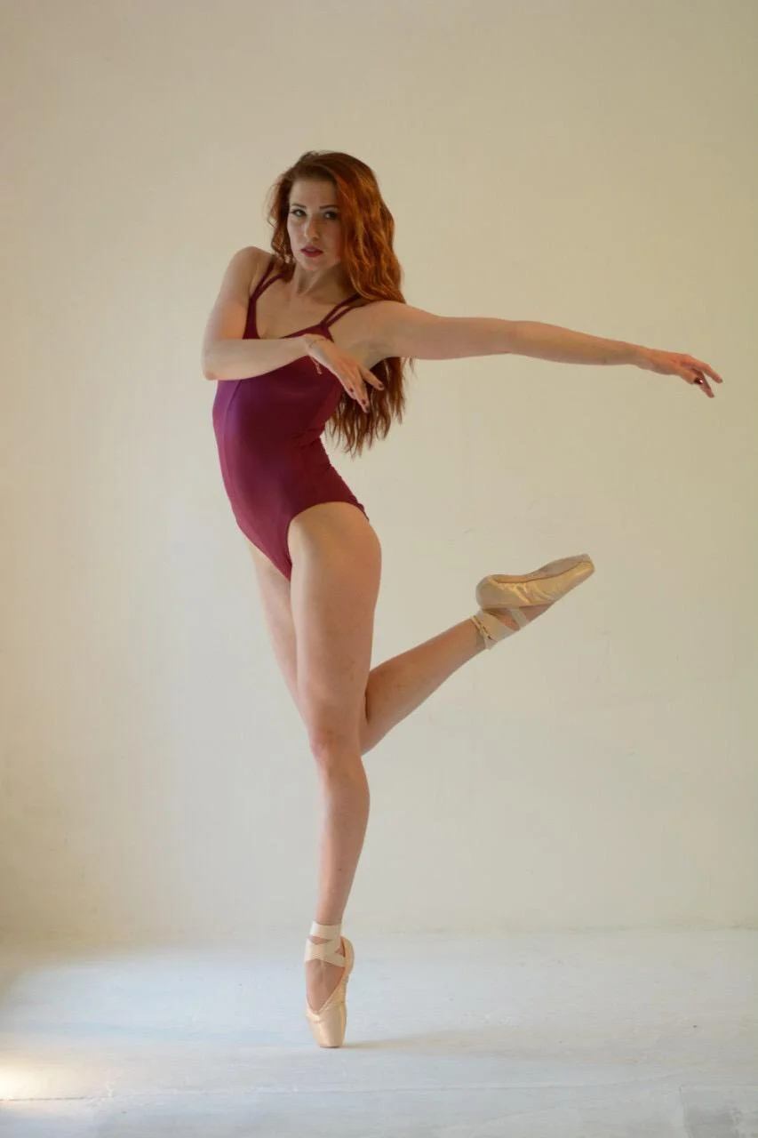 A ballet dancer with red hair wearing a burgundy leotard and pointe shoes, performing a pose with one leg raised and arms extended against a plain beige background.