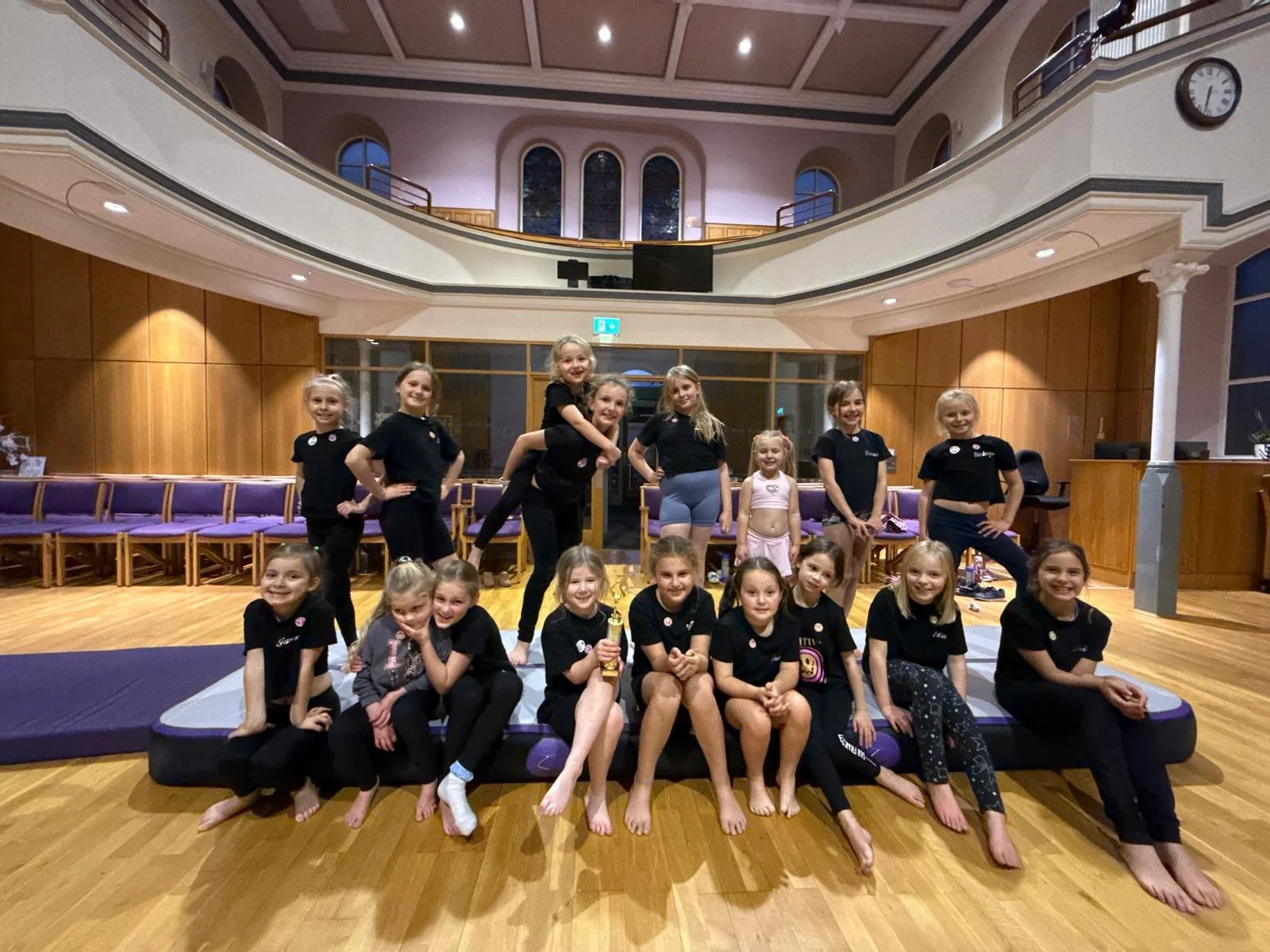 A group of young girls and women posing together in a spacious indoor hall with wooden floors and purple mats, some girls sitting and others standing, smiling at the camera.