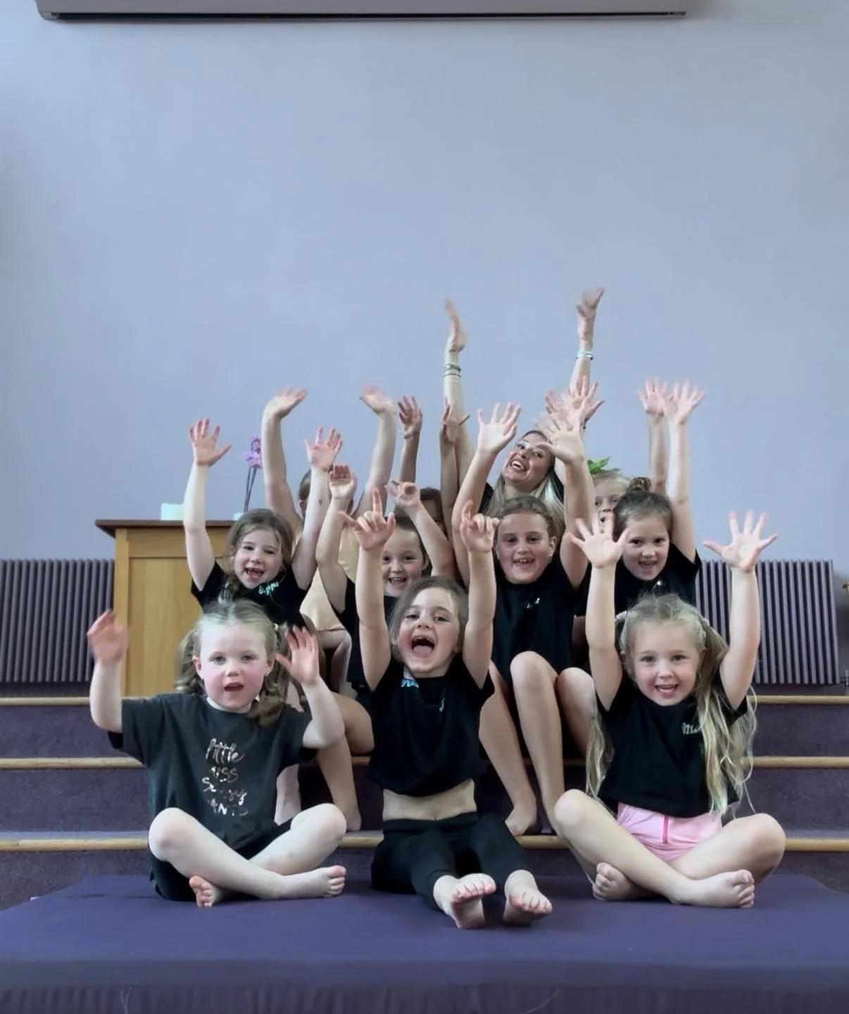 Group of children and a woman sitting on steps, smiling and raising their hands in a celebratory pose.