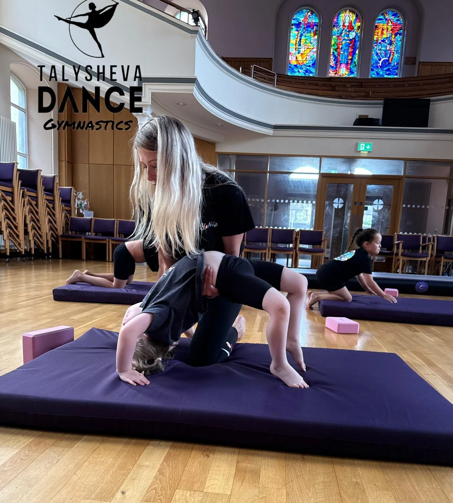 Young girl in a black shirt and shorts practicing gymnastics on a purple mat with an instructor in a gymnasium. Other children are seen practicing in the background. The gym has wooden floors, chairs, stained glass windows, and a balcony.