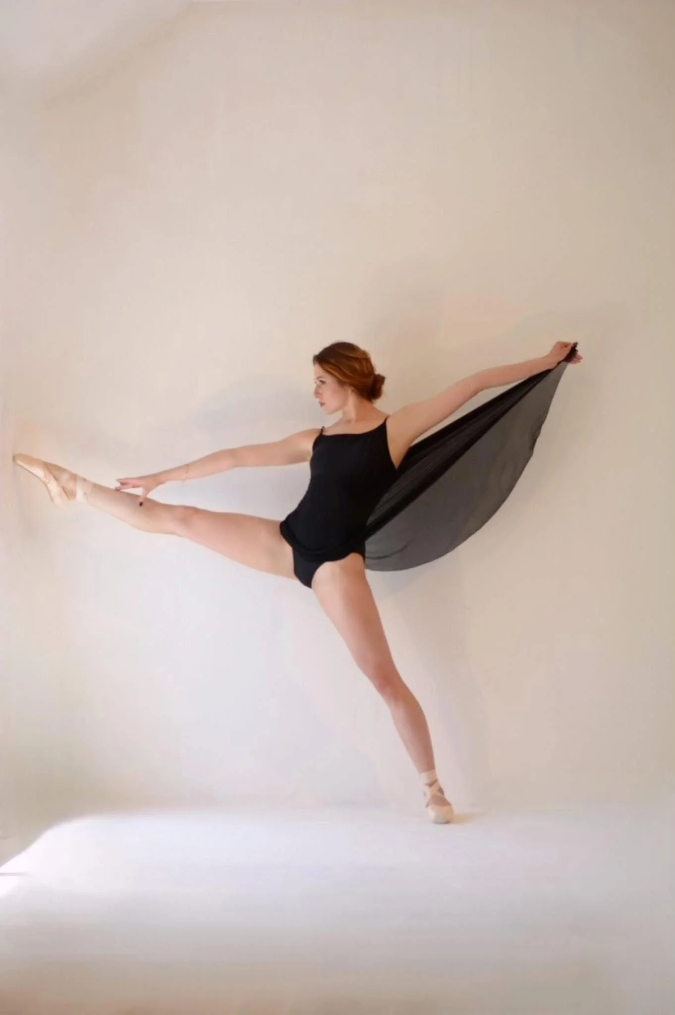 Ballet dancer in a black leotard and with a black flowing fabric performing a pose with one leg extended forward and arms stretched outward against a white background.