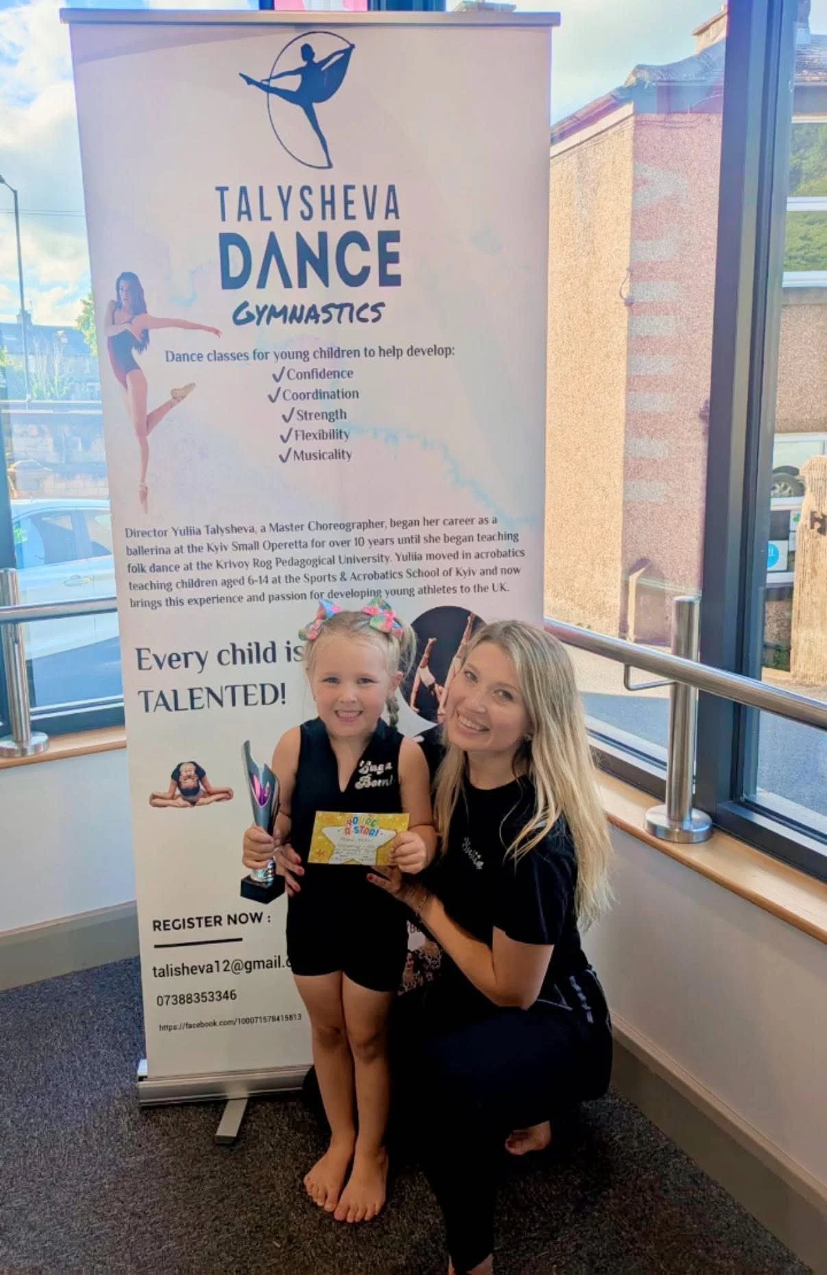 A young girl and an adult woman are smiling and posing for a photo indoors near a large banner for TALSHEVA DANCE GYMNASTICS. The girl is holding a colorful certificate and a small trophy. The banner promotes dance classes for children to develop con
