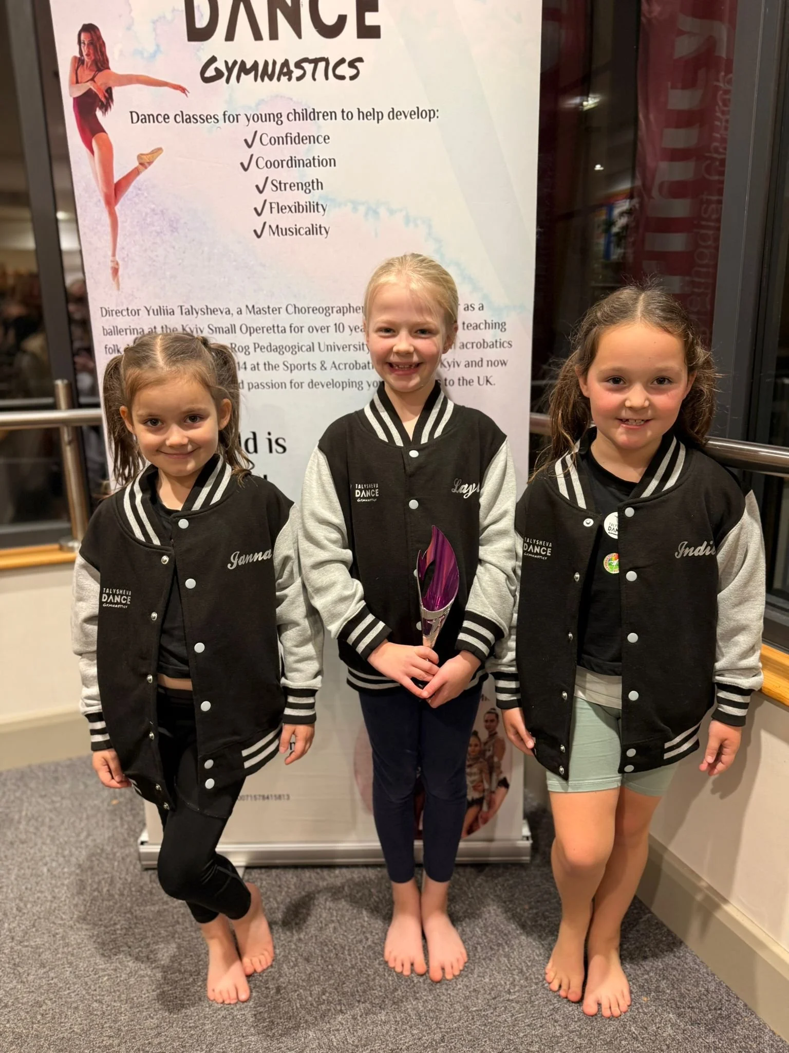 Three young girls standing barefoot indoors in front of a poster about dance gymnastics. They are wearing black and gray sports jackets with their names on the left chest: Janna, Layla, and Indira. Janna is on the left, Layla in the middle holding a 