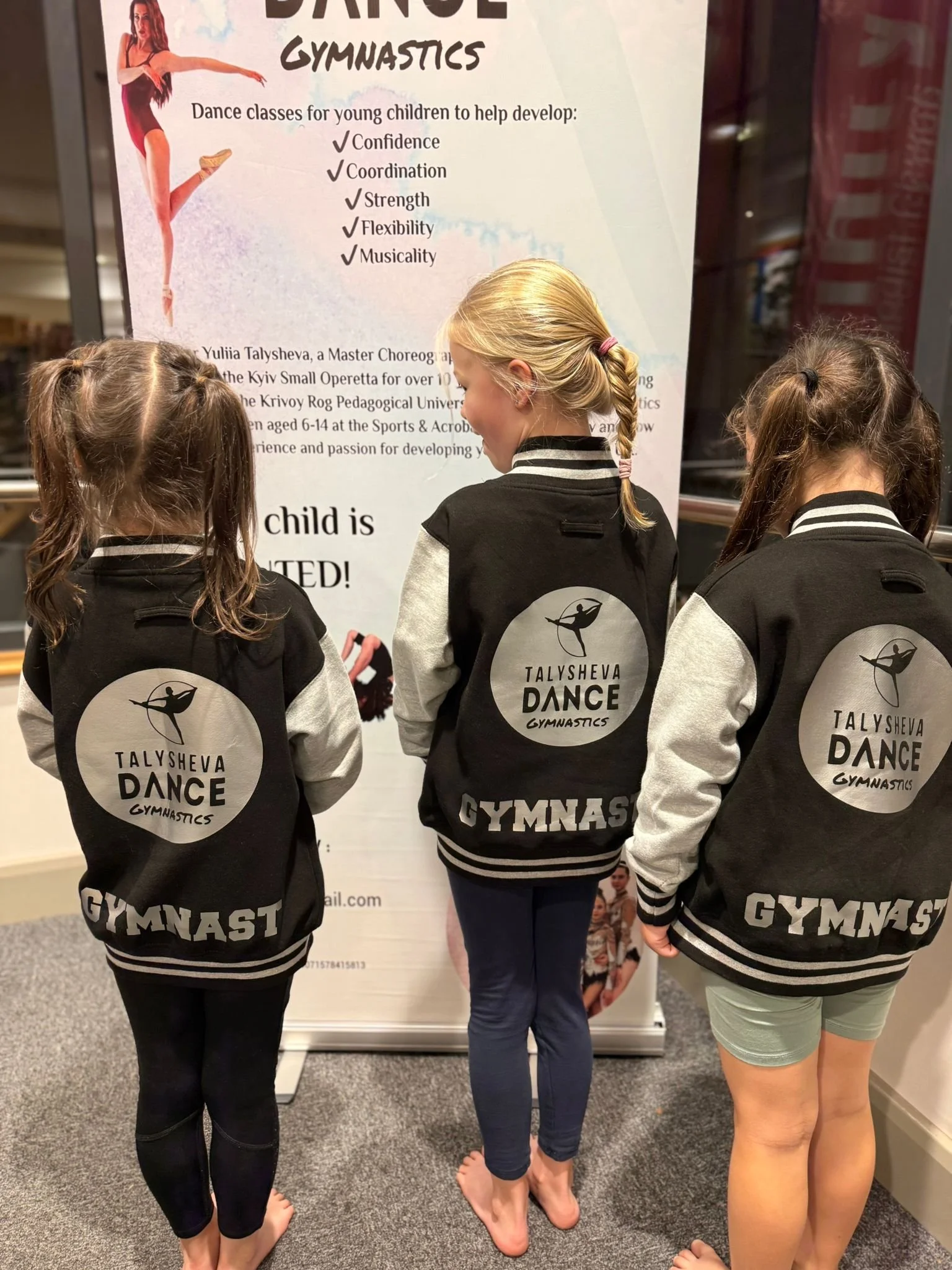 Three young girls with their backs to the camera, wearing black and gray varsity jackets with the logo and name of Talysheva Dance Gymnastics, standing in front of a poster advertising dance gymnastics classes for children.