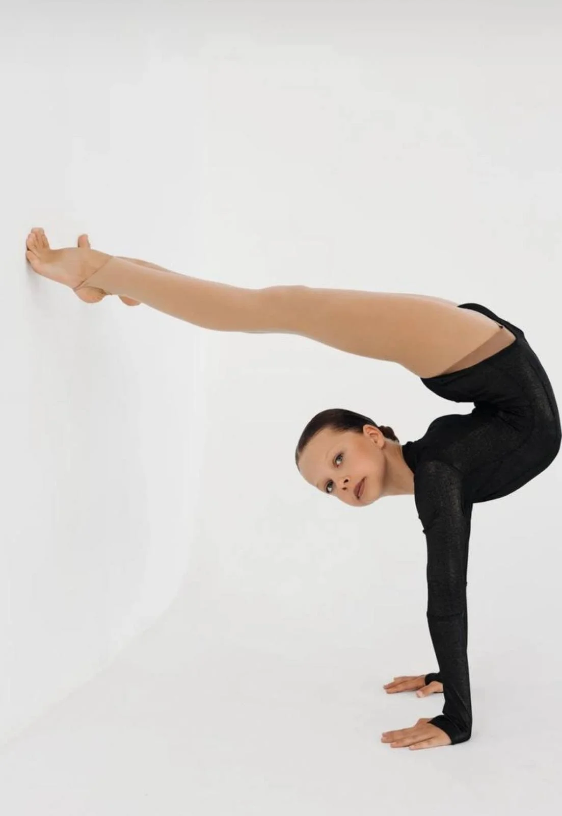 A young woman in black dance attire performing a handstand against a white wall, with her legs extended in a split position.