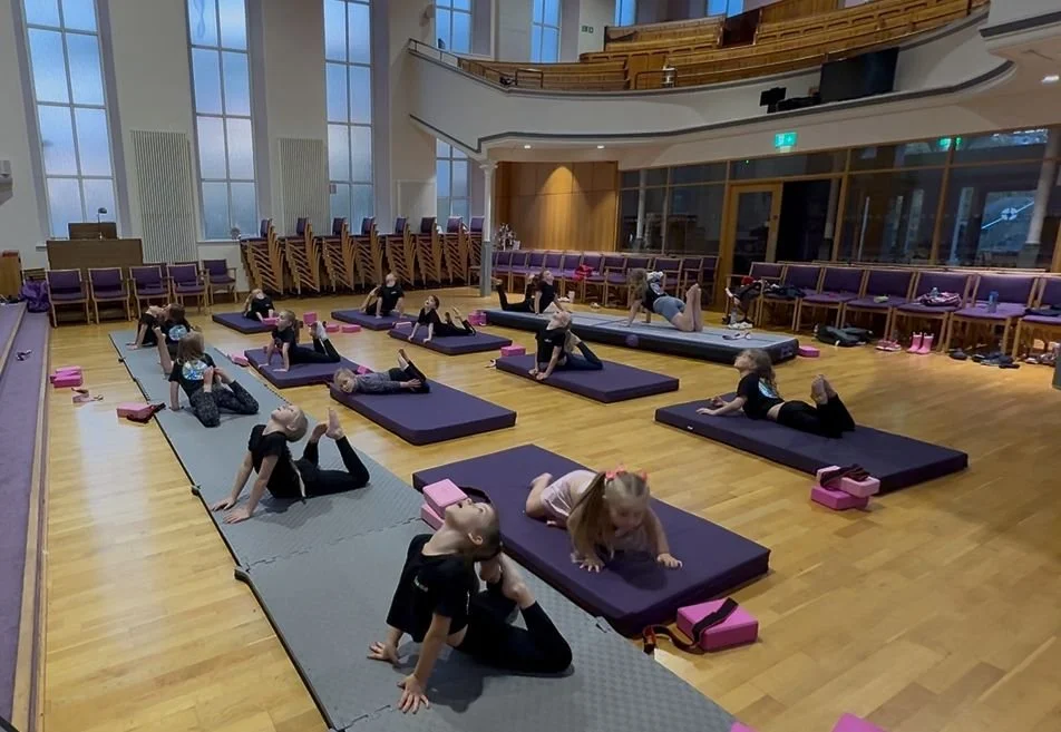 Children participating in a yoga class in a spacious indoor hall with large windows, black mats, pink yoga blocks, and a wooden floor.