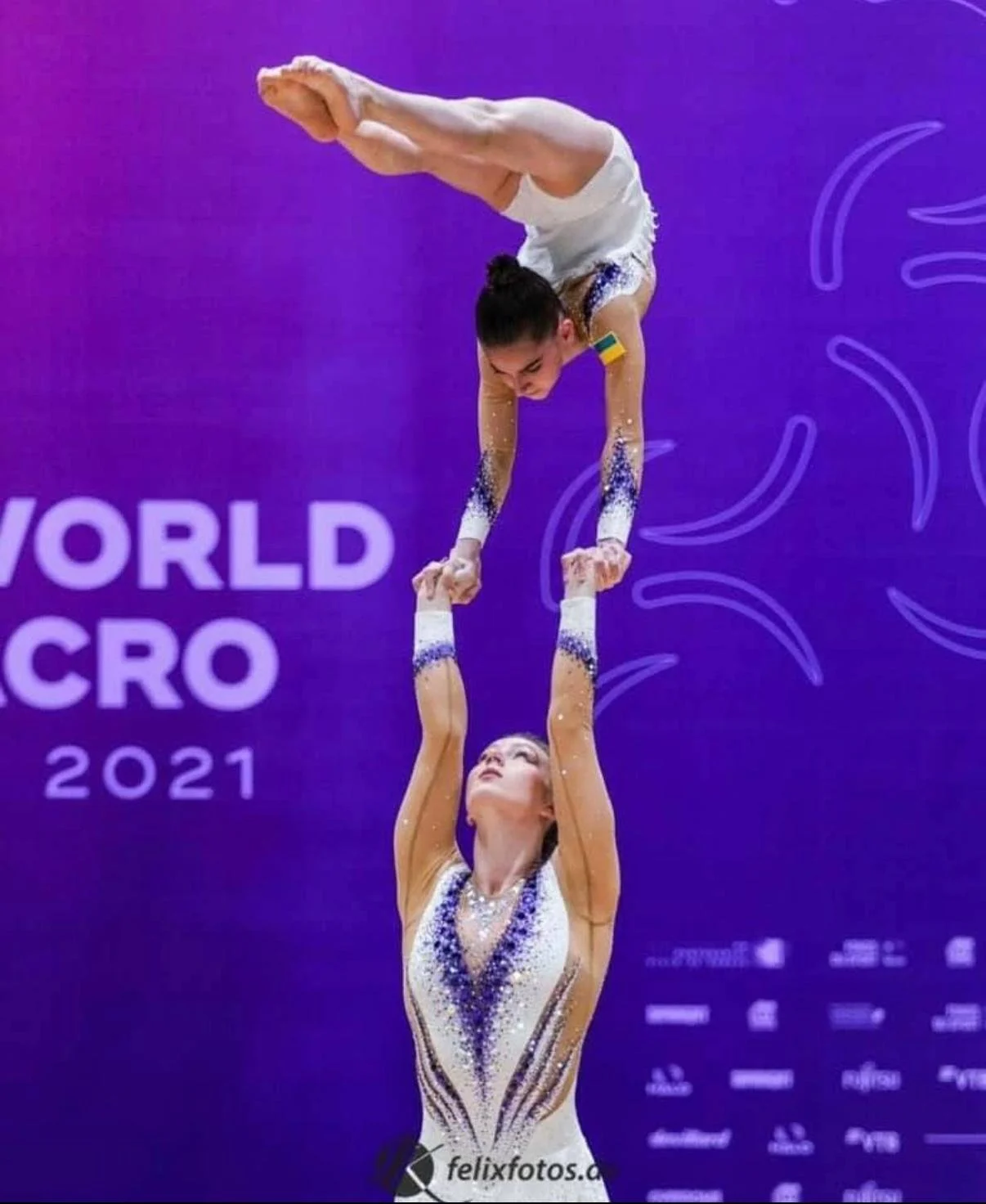 Two female acrobats performing a balancing act during a gymnastics competition with a purple backdrop. The woman on the ground is holding the other woman upside down by her hands.