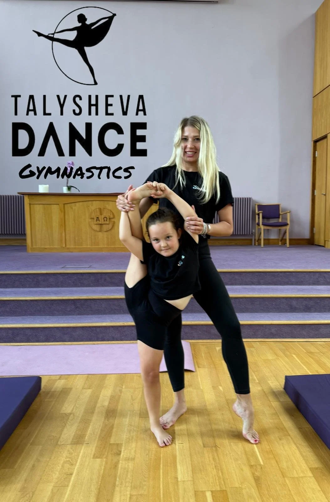 A woman and a young girl practicing gymnastics in a gymnasium, with a sign reading 'TALYSHEVA DANCE GYMNASTICS' on the wall behind them.