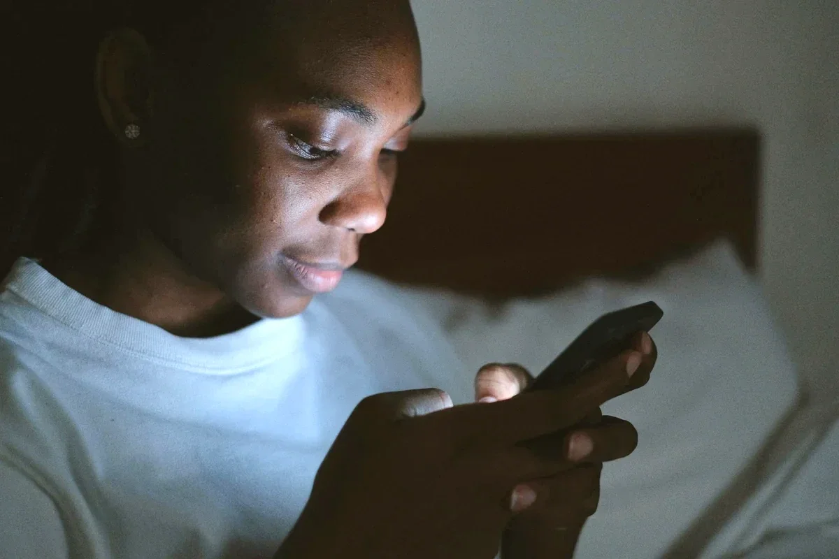 Person using a smartphone in a dark bedroom at night, face lit by blue screen glow before sleep.