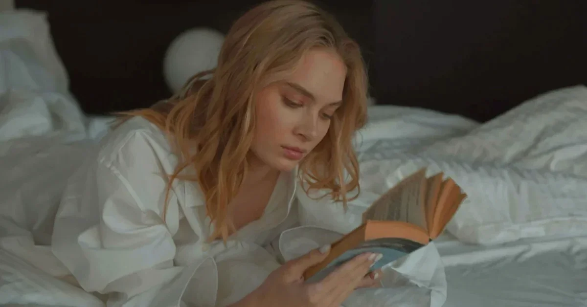 Woman lying in bed reading a book by warm lamp light, illustrating a calm screen-free bedtime routine.