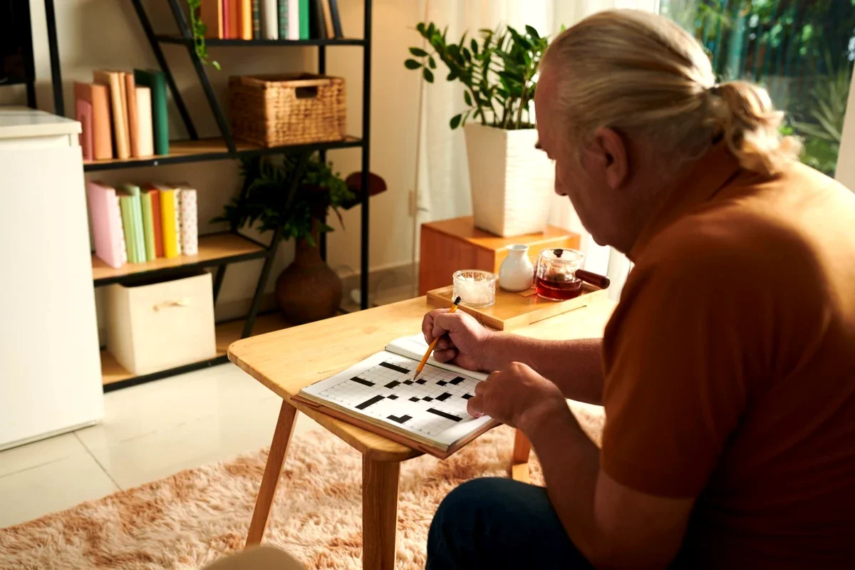 Senior man leaning over a wooden coffee table solving a crossword puzzle with a pencil in a warm, sunlit living room, illustrating the deep focus and calm concentration that puzzle solving provides.