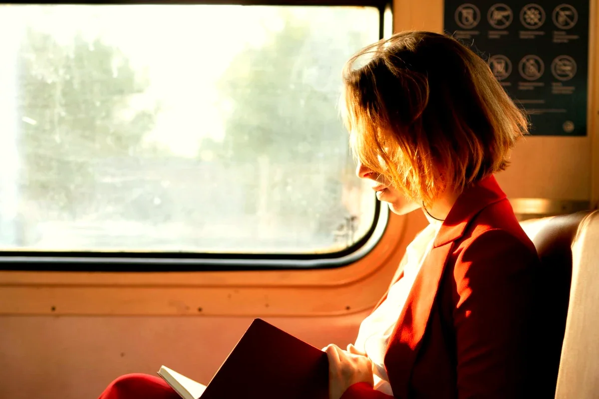 Woman in a red blazer reading a book by a train window bathed in warm golden sunlight, conveying the calm and focused mood of screen-free travel activity.