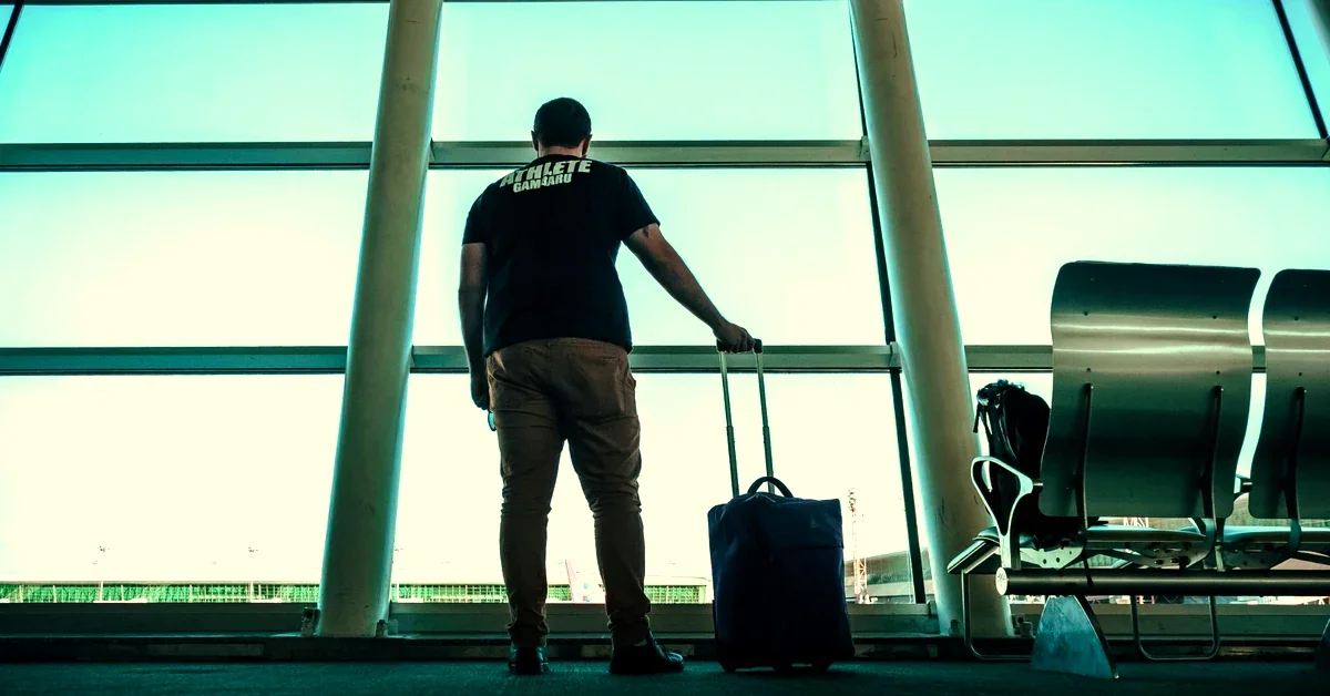 Traveller standing alone at an airport departure gate with a suitcase, looking out at the runway through large floor-to-ceiling windows, representing the quiet waiting time that puzzle books are perfect for.