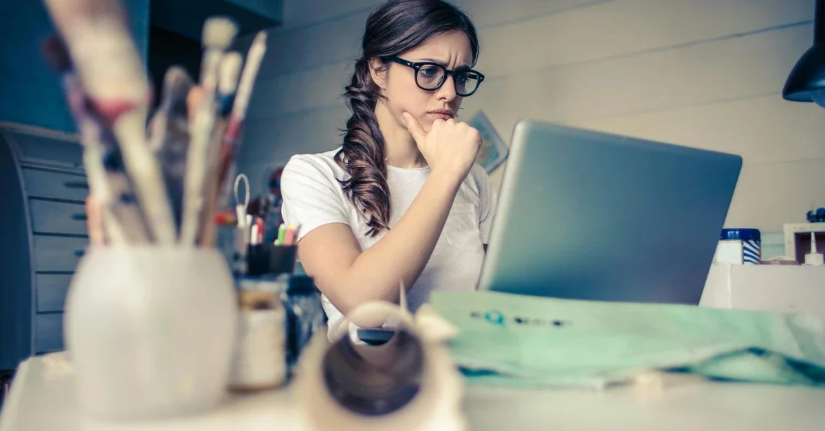 Young woman with glasses sitting at a desk, hand on chin, thinking deeply while looking at a laptop, representing the real-life problem-solving skills built through regular puzzle practice.