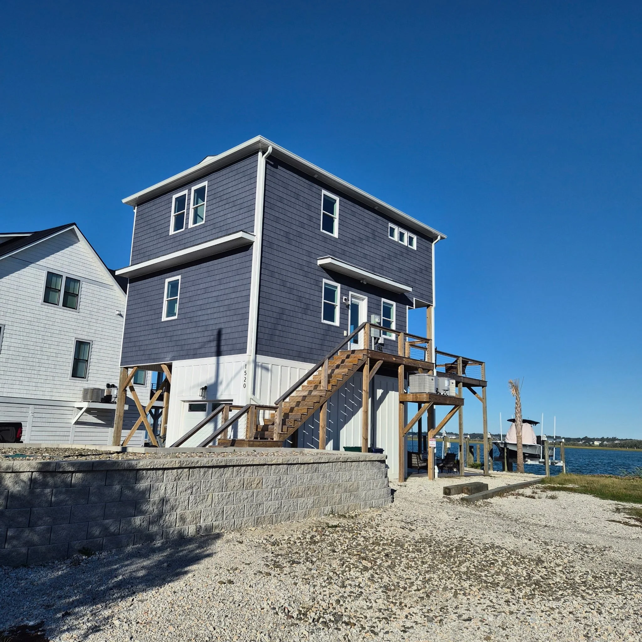 A modern three-story house with dark blue siding, white trim, and a wooden staircase leading to a deck. The house is elevated on stilts near a body of water with a boat docked in the background, under a clear blue sky.