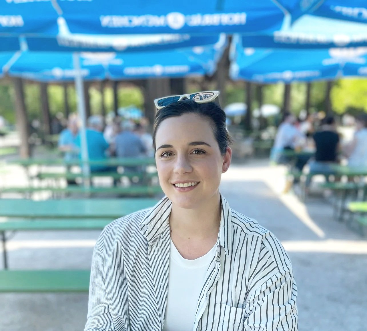 A smiling young woman with short dark hair and sunglasses on her head, wearing a striped shirt, sitting outdoors at a park with green benches and blue umbrellas in the background, during daytime.