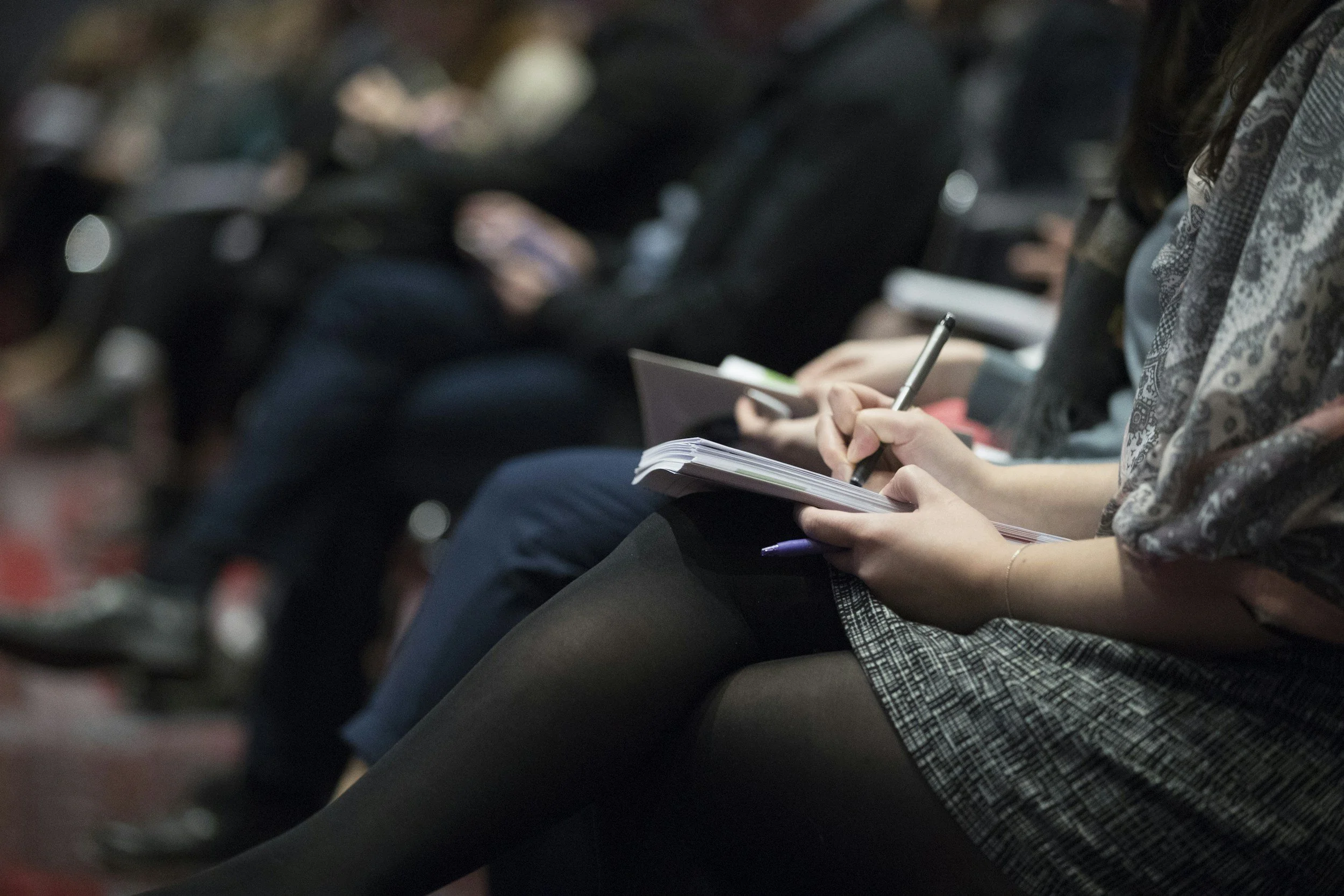 People sitting in an audience taking notes with pens and notebooks.