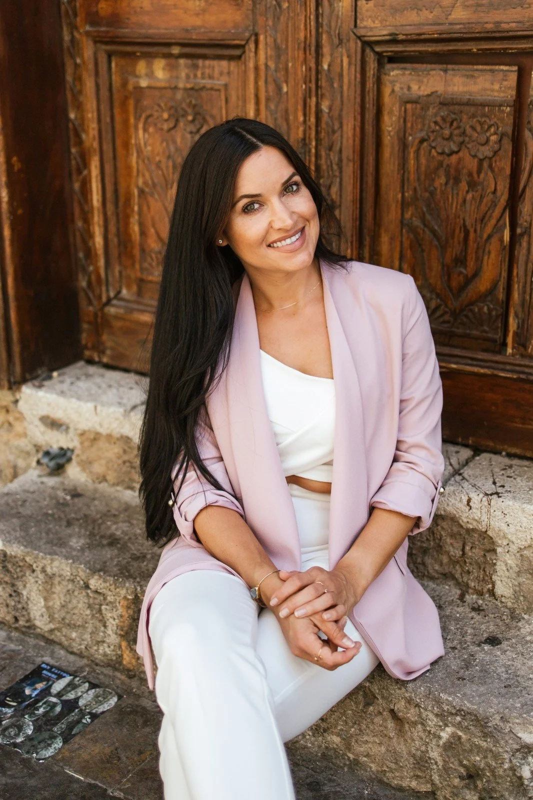 A woman with long dark hair, smiling, wearing a white top, light pink blazer, and white pants, sitting on stone steps in front of a wooden door with carved floral designs.