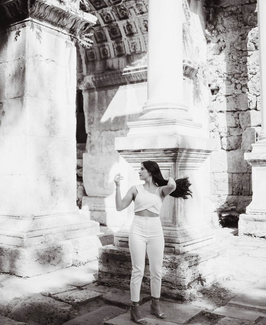 A woman with dark hair in a light-colored crop top and high-waisted pants standing among ancient stone ruins with Corinthian columns