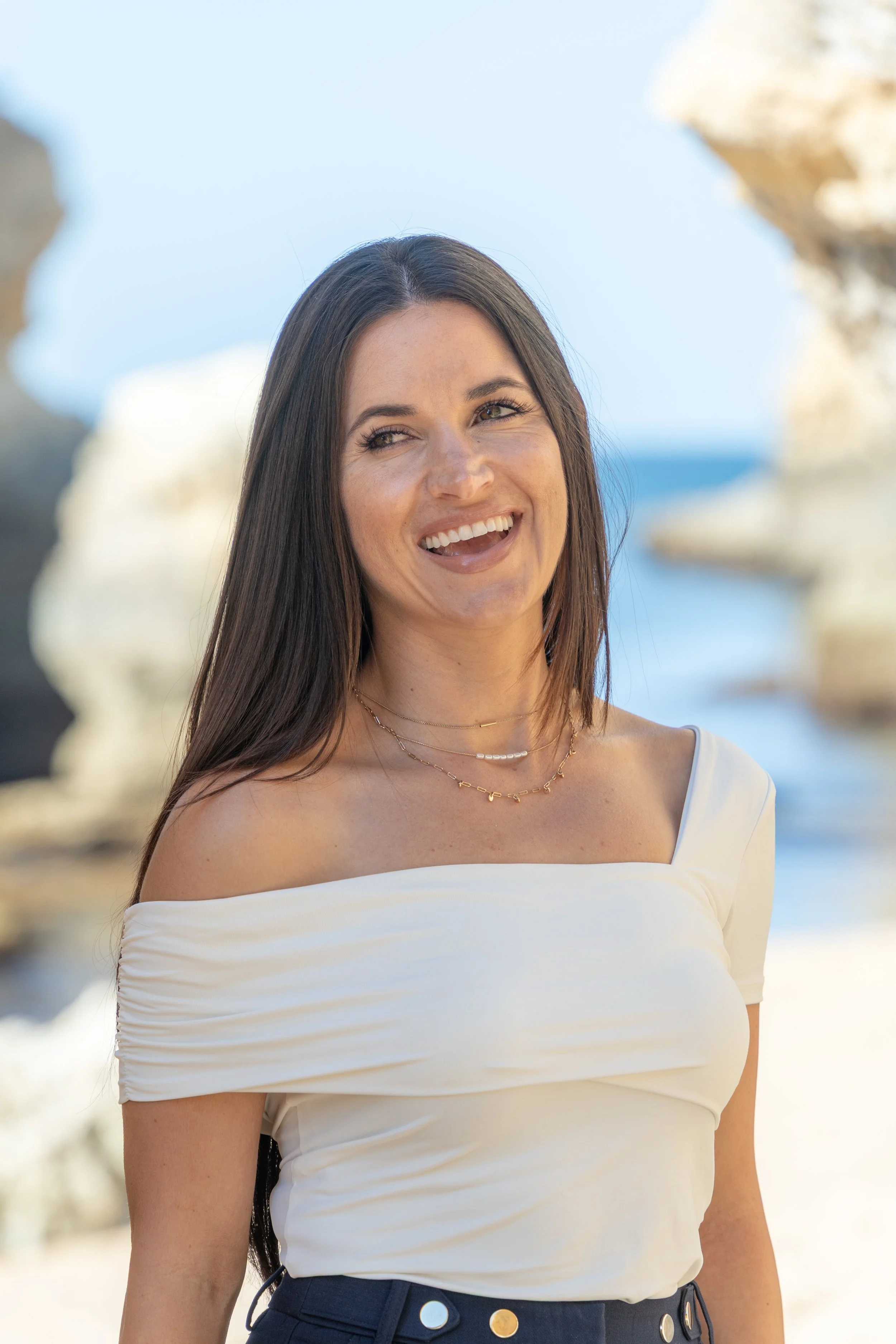A woman with long brown hair smiling outdoors with large rock formations and a blue sky in the background.