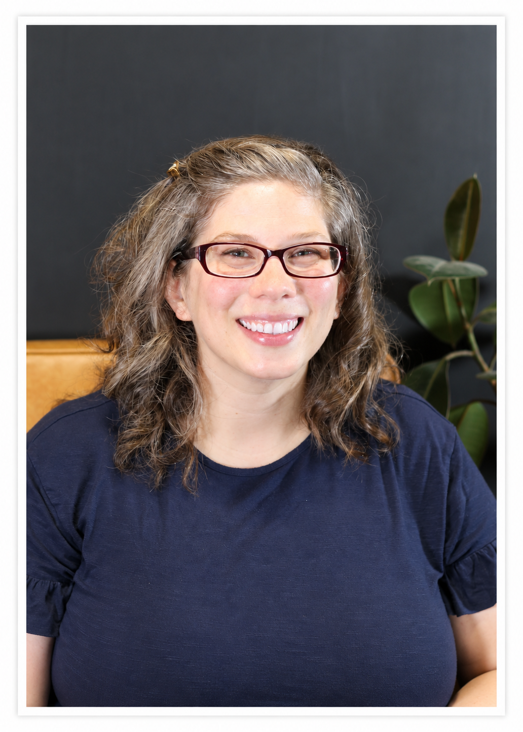 A smiling woman with glasses and curly hair sitting in front of a dark background and a potted plant.