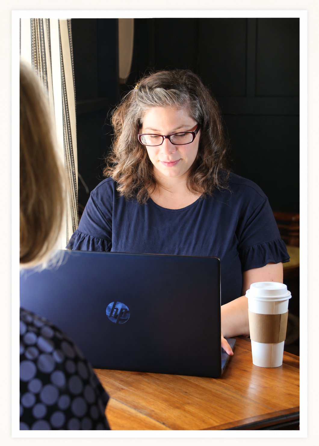 A woman with glasses and wavy hair working on a laptop at a wooden table, with a paper coffee cup nearby, in a cozy indoor setting.