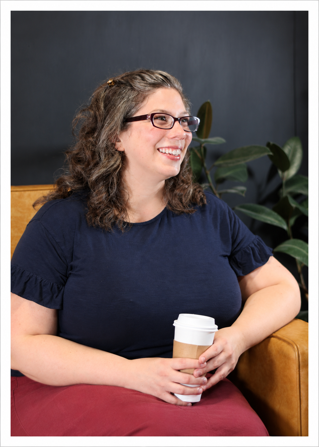 A smiling woman with glasses and curly hair sitting on a wooden chair, holding a disposable coffee cup. She wears a navy blue shirt with ruffled sleeves and red pants, with green houseplants in the background.