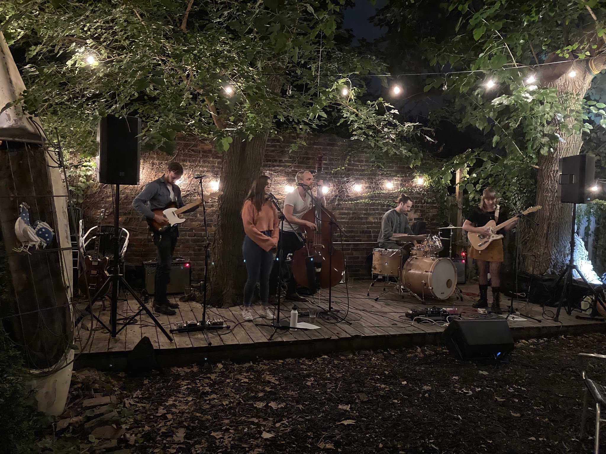 A band performing live on an outdoor stage at night, surrounded by trees and string lights, with five musicians playing guitars, drums, and singing.