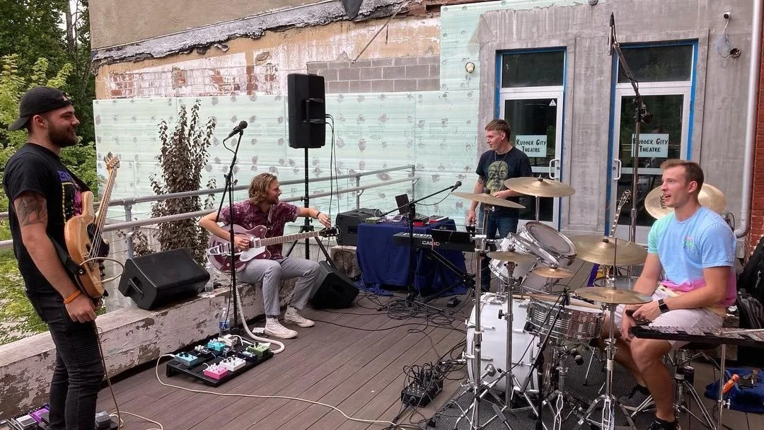 A band performing outdoors on a wooden stage, with four members playing instruments and a sound system set up. The background shows an unfinished brick wall with a window and door.