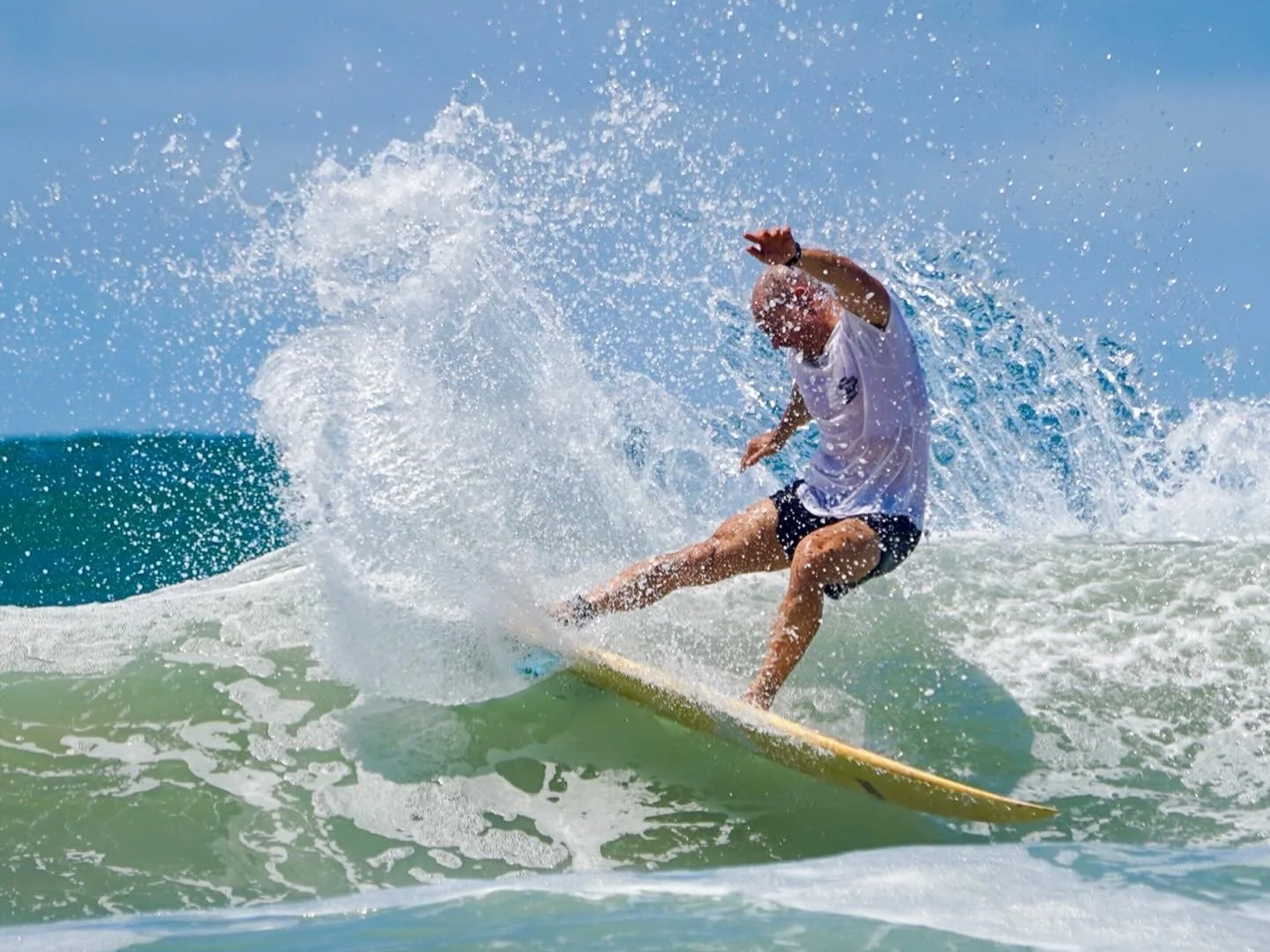 A person surfing on a wave in the ocean, wearing a white T-shirt and black shorts, with water splashing around.