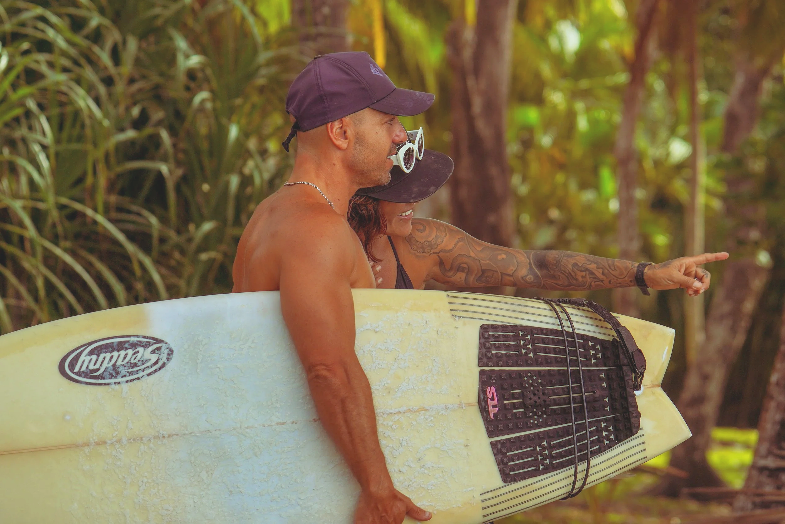 Two surfers, a man and a woman, standing together in a tropical forest, with the woman pointing at something in the distance. They are holding a surfboard with a guitar neck and keys attached to it, and wearing hats and sunglasses.